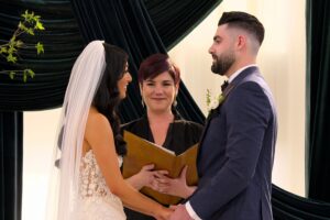 A woman in a wedding dress and a man in a suit face eachother and hold hands at a wedding alter. A person holding an open folder stands behind them.