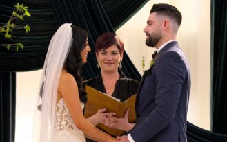 A woman in a wedding dress and a man in a suit face eachother and hold hands at a wedding alter. A person holding an open folder stands behind them.