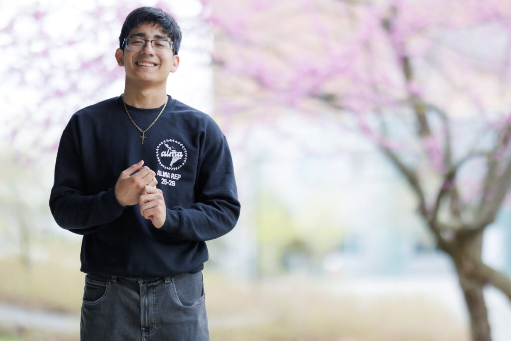 Paul Vargas is outside on campus and stands left of center, smiling with clasped hands. There is a purple flowering tree in the background.