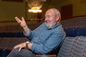 A man sits in a seat inside a theater, smiles and gestures forward with his hand.