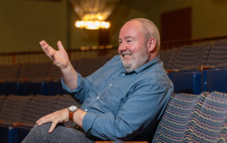 A man sits in a seat inside a theater, smiles and gestures forward with his hand.