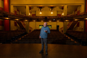 A man stands on stage in a theater, with empty seats behind him.