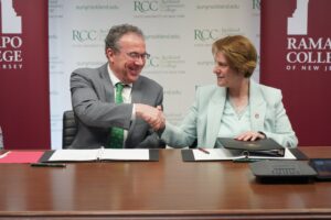 RCC President William Mullaney is on the left shaking hands with RCNJ President Cindy Jebb, who is on the right. Both are seated at a table with folios containing the signed transfer agreement.