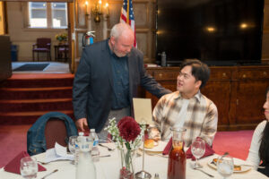 A man stands and puts his hand on the shoulder of a young man sitting at a table.