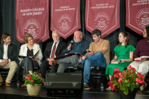Seven people sit on chairs on a stage, with maroon banners behind them. The man in the middle talks and holds a microphone.