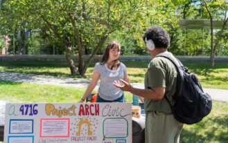 Two students are outside talking at a table in the student involvement fair. There is a colorful poster on the table that describes the student club "Project ARCH."