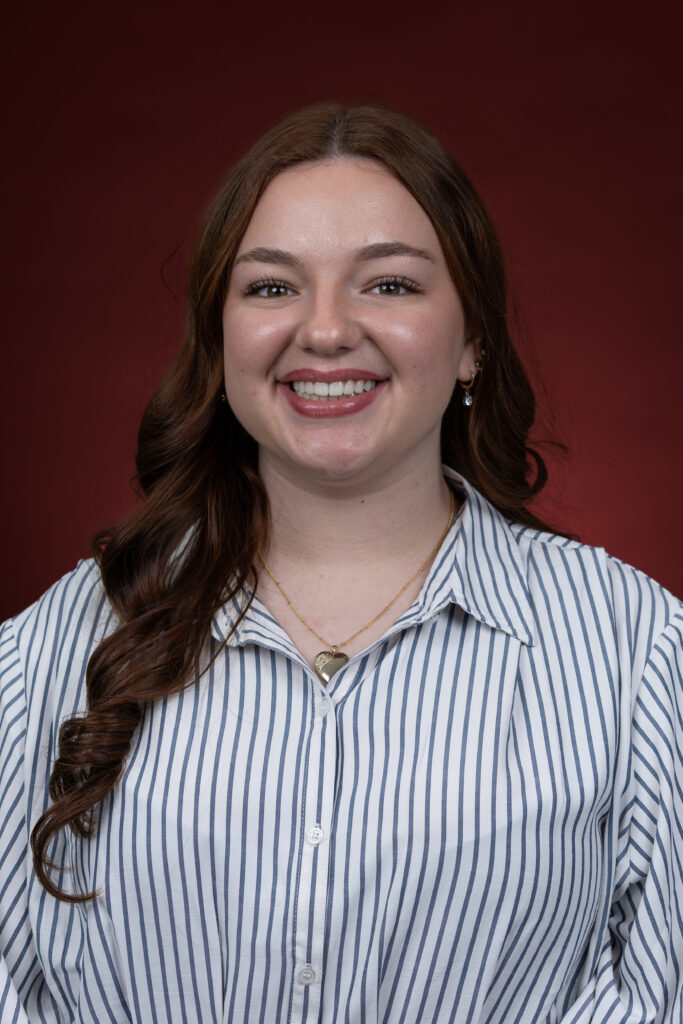 Sarah is facing the camera, smiling. Her long hair cascades over her right shoulder. She is wearing a striped button-down shirt and is standing in front of a maroon backdrop.