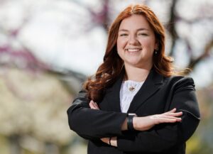 Sarah Glisson stands outside in a black suit, smiling with her arms crossed.