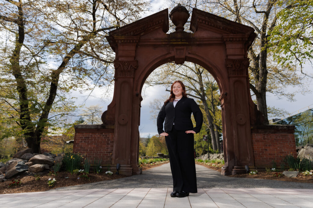 Sarah Glisson is standing in front of the Havemeyer Arch on campus. She is wearing a black suit and has her hands on her hips. The camera angle is looking up at her.