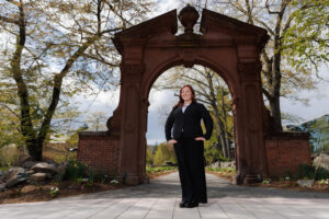 Sarah Glisson is standing in front of the Havemeyer Arch on campus. She is wearing a black suit and has her hands on her hips. The camera angle is looking up at her.