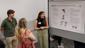 A young woman stands in front of a poster and talks to two people.
