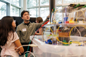 A male college student points to a research paper and speaks to a young woman. A STEM project is in front of him.