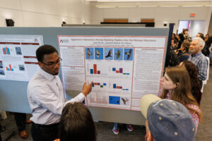 A student stands in front of a research poster and speaks as others look on.