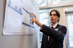 A college student in a black suit points at a research poster and speaks.
