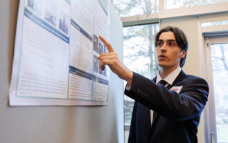 A college student in a black suit points at a research poster and speaks.