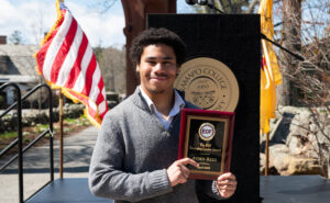 A young man stands outside holding a plaque. He is smiling. 