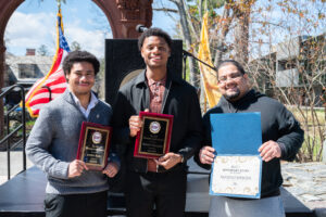 Three young men stand outside smiling and holding plaques.