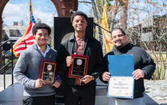Three young men stand outside smiling and holding plaques.