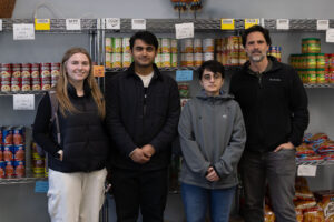 A group of four people stand together in front of shelves of food in a food pantry.