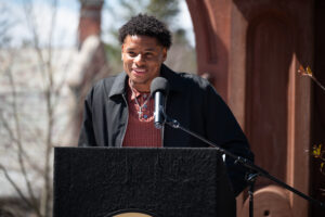 A young man leans over a podium and speaks. He is outside.