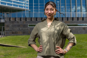 A woman stands outside in front of a glass building. She poses with her hands on her hips.