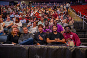 Five people sit in the front row of an arena. A crowd fills the seats behind them.