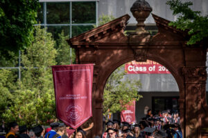 A person in a cap and gown carries a large maroon gonfalon through a large brick arch. She is surrounded by other people in graduation gowns.