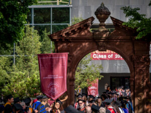 A person in a cap and gown carries a large maroon gonfalon through a large brick arch. She is surrounded by other people in graduation gowns.