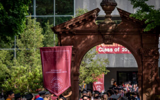 A person in a cap and gown carries a large maroon gonfalon through a large brick arch. She is surrounded by other people in graduation gowns.