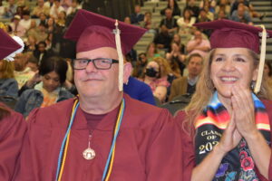 A man in a maroon cap and gown sits smiling, while a woman in a maroon cap and gown and colorful stole smiles and claps next to him.