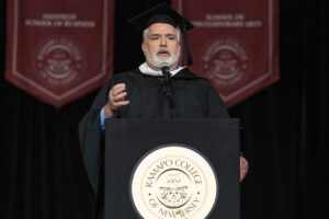 A man wearing a black graduation gown and cap stands behind a podium wiht a gold seal on it and speaks.