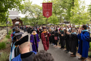 A man in a maroon graduation cap and gown holds a gonfolan and walks down a road. A woman in a purple and black cap and gown walks beside him. The road is lined with people cheering.
