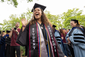 A woman in a black cap and gown smiles wide outside.