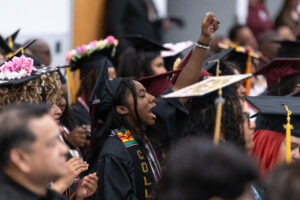 A woman in a black graduation gown sits in a crowd of graduates with her fist up and her mouth open. 