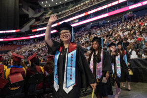 A woman in a graduation gown and hat looks up, waves and smiles. She is in a crowded arena.