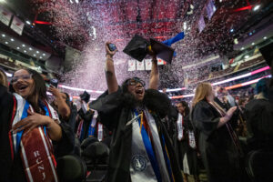 A woman in a graduation gown holds up her cap and her hand in the air as confetti falls inside an arena.