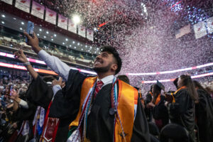 A graduate in a black gown and yellow stole looks up and smiles as confetti pours down inside an arena. His arm is stretched up.
