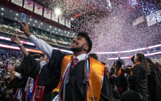 A graduate in a black gown and yellow stole looks up and smiles as confetti pours down inside an arena. His arm is stretched up.