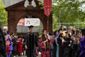 A person in a maroon graduation robe and cap carries a gonfolan, and walks next to a man in a black gown and cap. They are outside in front of a large arch. They are surrounded by others in caps and gowns.