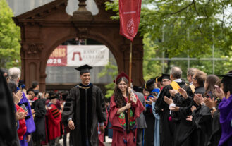 A person in a maroon graduation robe and cap carries a gonfolan, and walks next to a man in a black gown and cap. They are outside in front of a large arch. They are surrounded by others in caps and gowns.