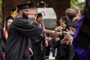 A student in a black cap and gown shakes hands with someone in a crowd outside in front of an arch.