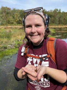 A woman in a maroon shirt stands outside holding a newt in her hands. She is in front of a pond and wearing a backpack.
