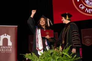 A woman in a black graduation robe and cap holds a diploma and her other hand in the air, as another woman in graduation gown and cap smiles at her.