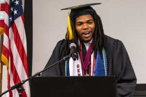 A young man in a black graduation cap and gown stands at a podium inside and speakss. A microphone is in front of him.