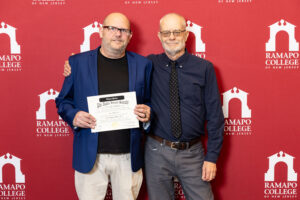 Two men stand next to eachother in front of a maroon wall with white arches and the words Ramapo Colelge. One is holding a certificate.