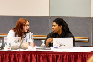 Two college students sit at a table and talk. Laptops are in front of them.