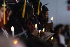 Students in black caps and gowns hold candles indoors. They have yellow tassels hanging from their caps.