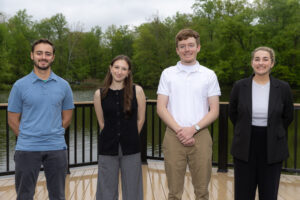 Four college students stand next to eachother on a dock, with a pond behind them abd trees behind the pond.