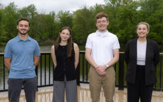 Four college students stand next to eachother on a dock, with a pond behind them abd trees behind the pond.