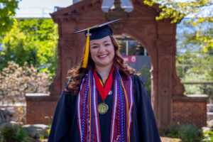 A woman in a graduation cap and gown stands in front of a brick arch outdoors.